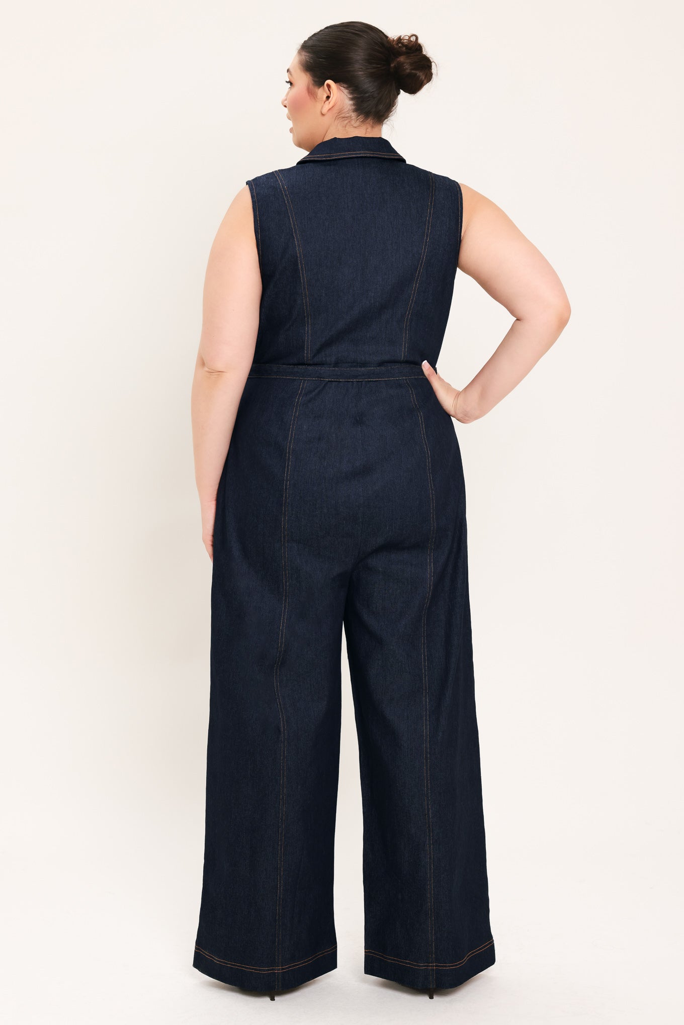 Woman wearing a dark blue denim sleeveless, wide-leg jumpsuit featuring collar, front zipper closure, and pockets | Plus Curve  FREEMARKET