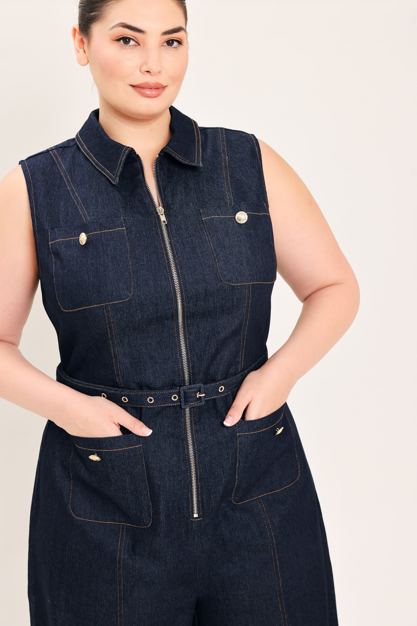 Woman wearing a dark blue denim sleeveless, wide-leg jumpsuit featuring collar, front zipper closure, and pockets | Plus Curve  FREEMARKET