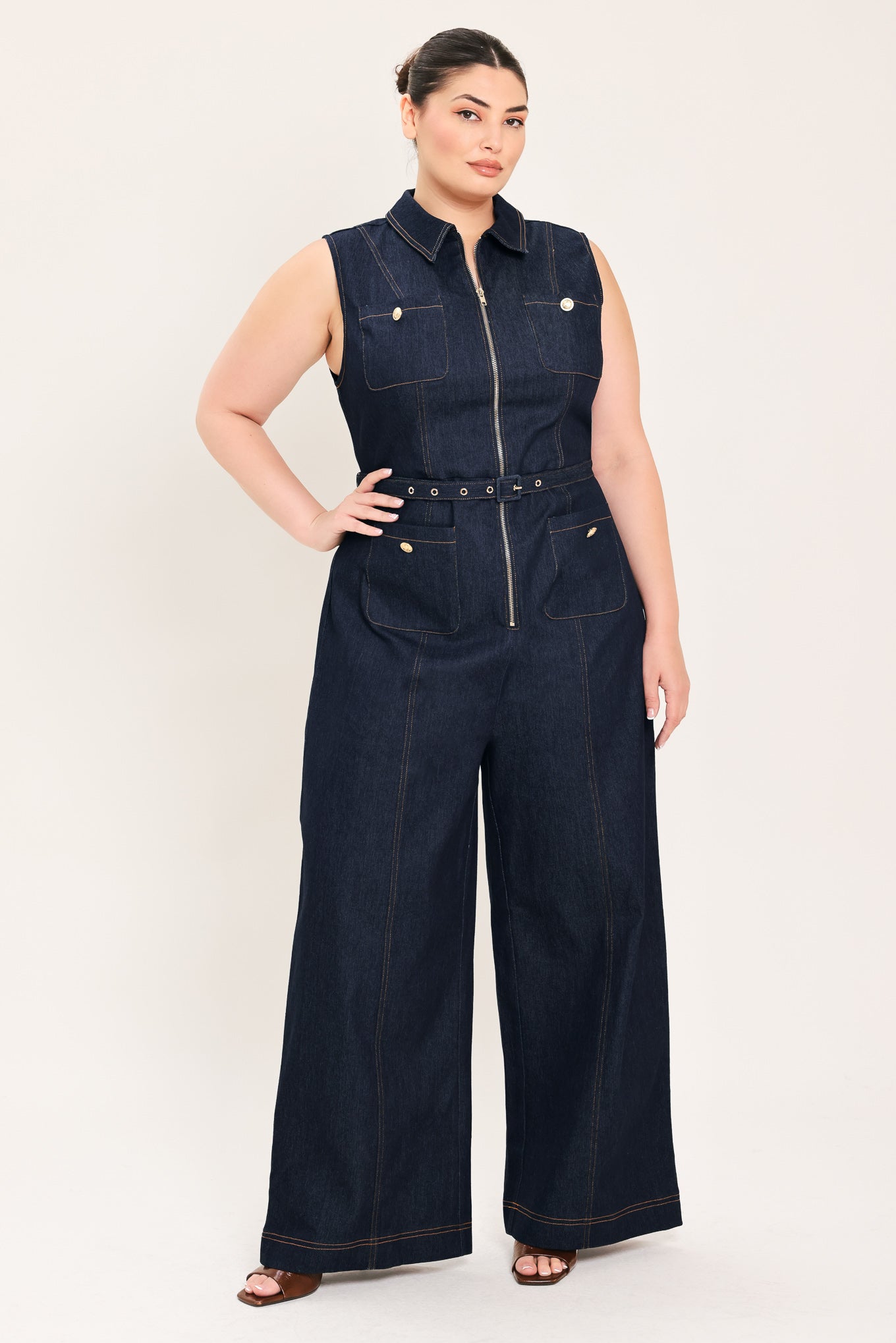 Woman wearing a dark blue denim sleeveless, wide-leg jumpsuit featuring collar, front zipper closure, and pockets | Plus Curve  FREEMARKET