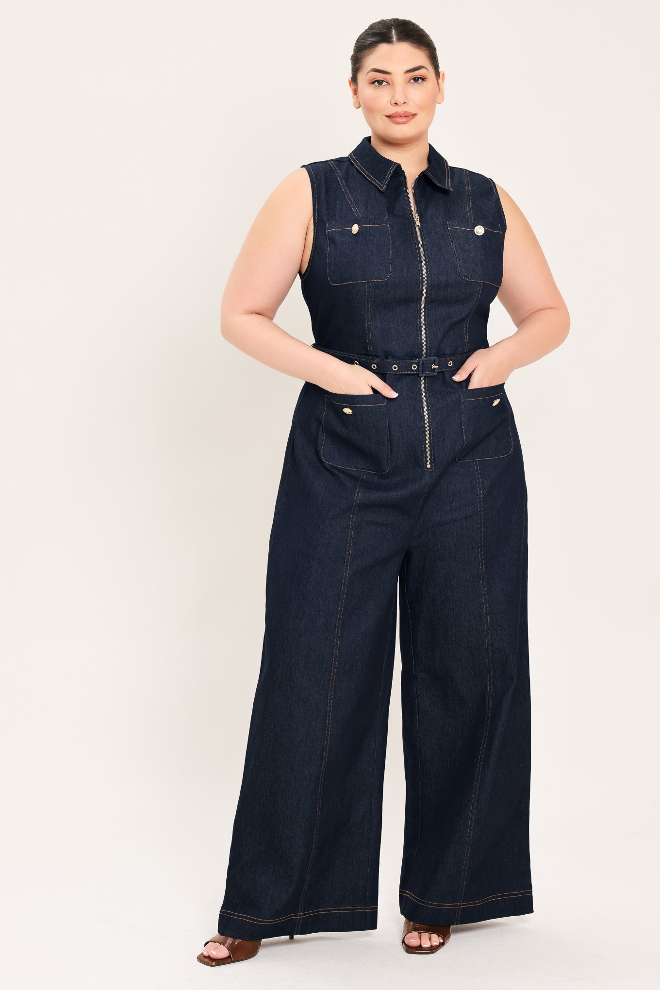 Woman wearing a dark blue denim sleeveless, wide-leg jumpsuit featuring collar, front zipper closure, and pockets | Plus Curve  FREEMARKET