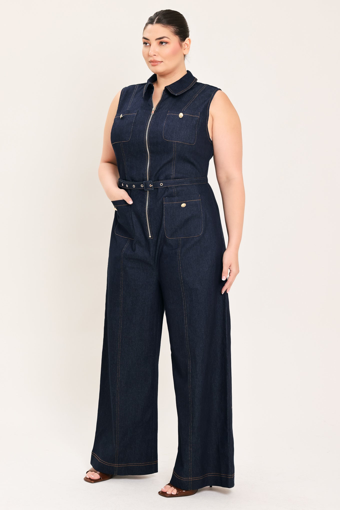 Woman wearing a dark blue denim sleeveless, wide-leg jumpsuit featuring collar, front zipper closure, and pockets | Plus Curve  FREEMARKET