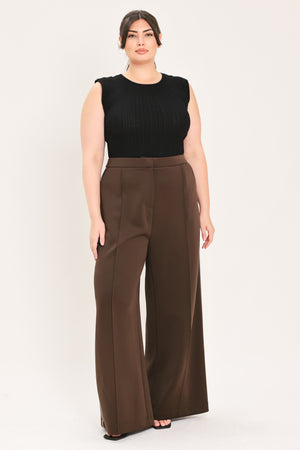 A woman modeling a sleeveless black ribbed top and high-waisted brown wide-leg trousers against a plain light gray studio background.
