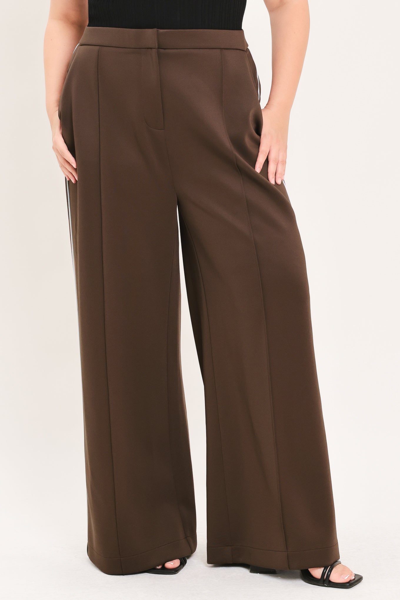 A model wearing high-waisted, wide-leg brown trousers with a black top and black sandals, shown from the waist down against a light beige studio background.