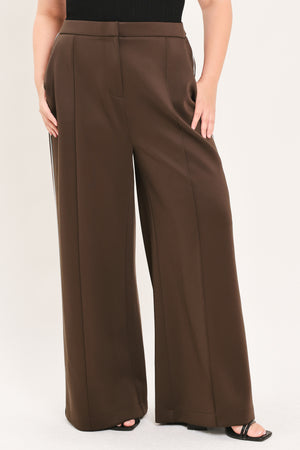A model wearing high-waisted, wide-leg brown trousers with a black top and black sandals, shown from the waist down against a light beige studio background.