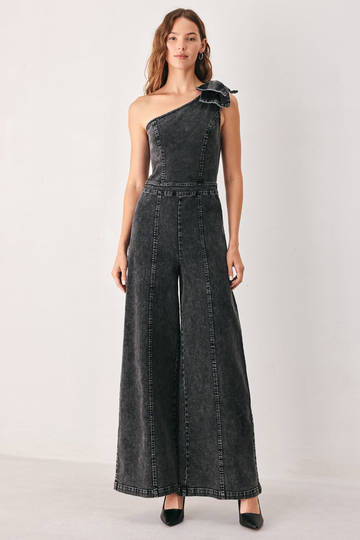 A woman wearing a dark gray washed denim one-shoulder jumpsuit with a wide-leg pant leg and a ruffled bow detail on the shoulder | FREEMARKET