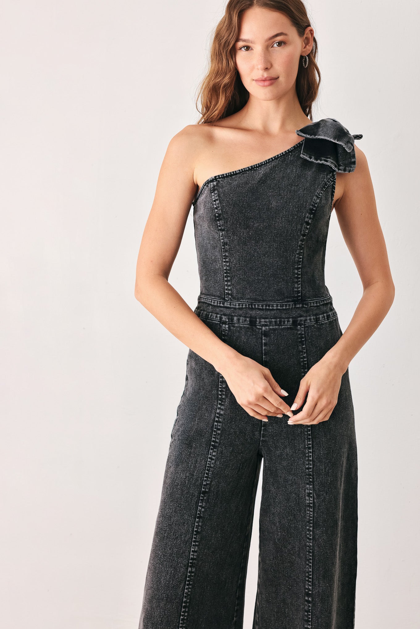 A woman wearing a dark gray washed denim one-shoulder jumpsuit with a wide-leg pant leg and a ruffled bow detail on the shoulder | FREEMARKET