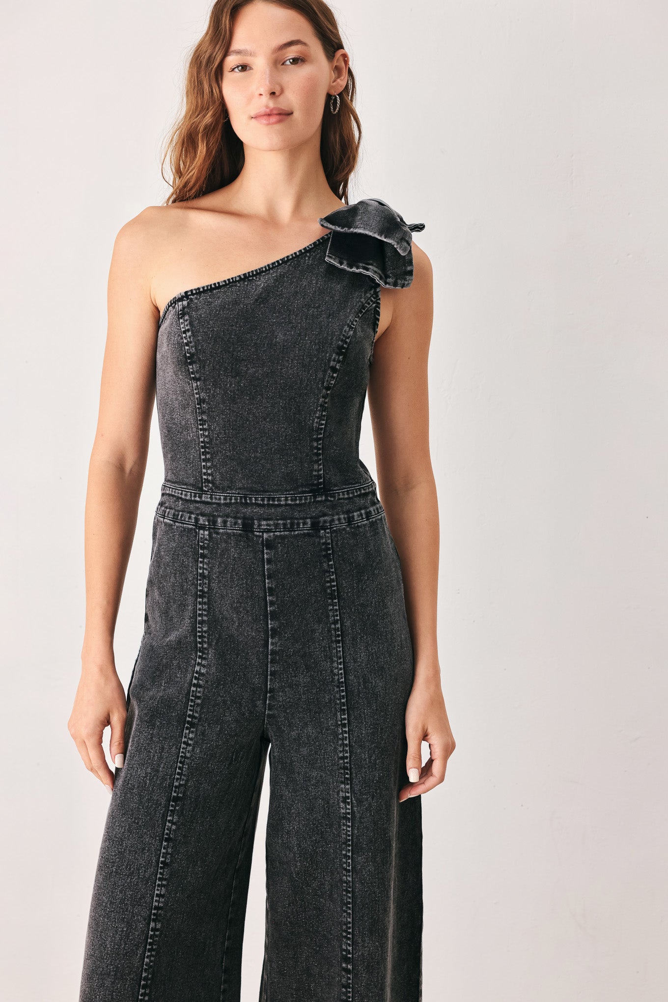 A woman wearing a dark gray washed denim one-shoulder jumpsuit with a wide-leg pant leg and a ruffled bow detail on the shoulder | FREEMARKET