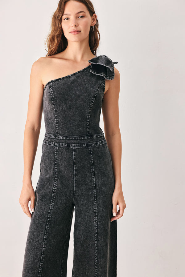 A woman wearing a dark gray washed denim one-shoulder jumpsuit with a wide-leg pant leg and a ruffled bow detail on the shoulder | FREEMARKET