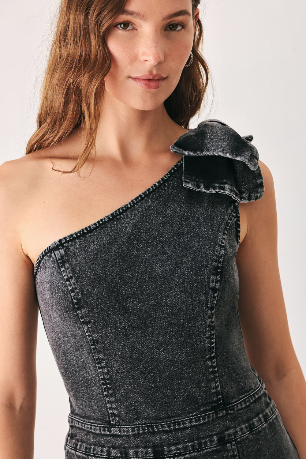 A woman wearing a dark gray washed denim one-shoulder jumpsuit with a wide-leg pant leg and a ruffled bow detail on the shoulder | FREEMARKET