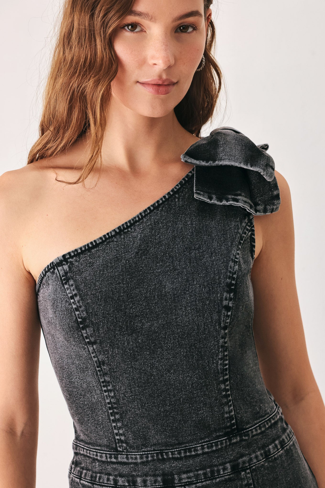 A woman wearing a dark gray washed denim one-shoulder jumpsuit with a wide-leg pant leg and a ruffled bow detail on the shoulder | FREEMARKET