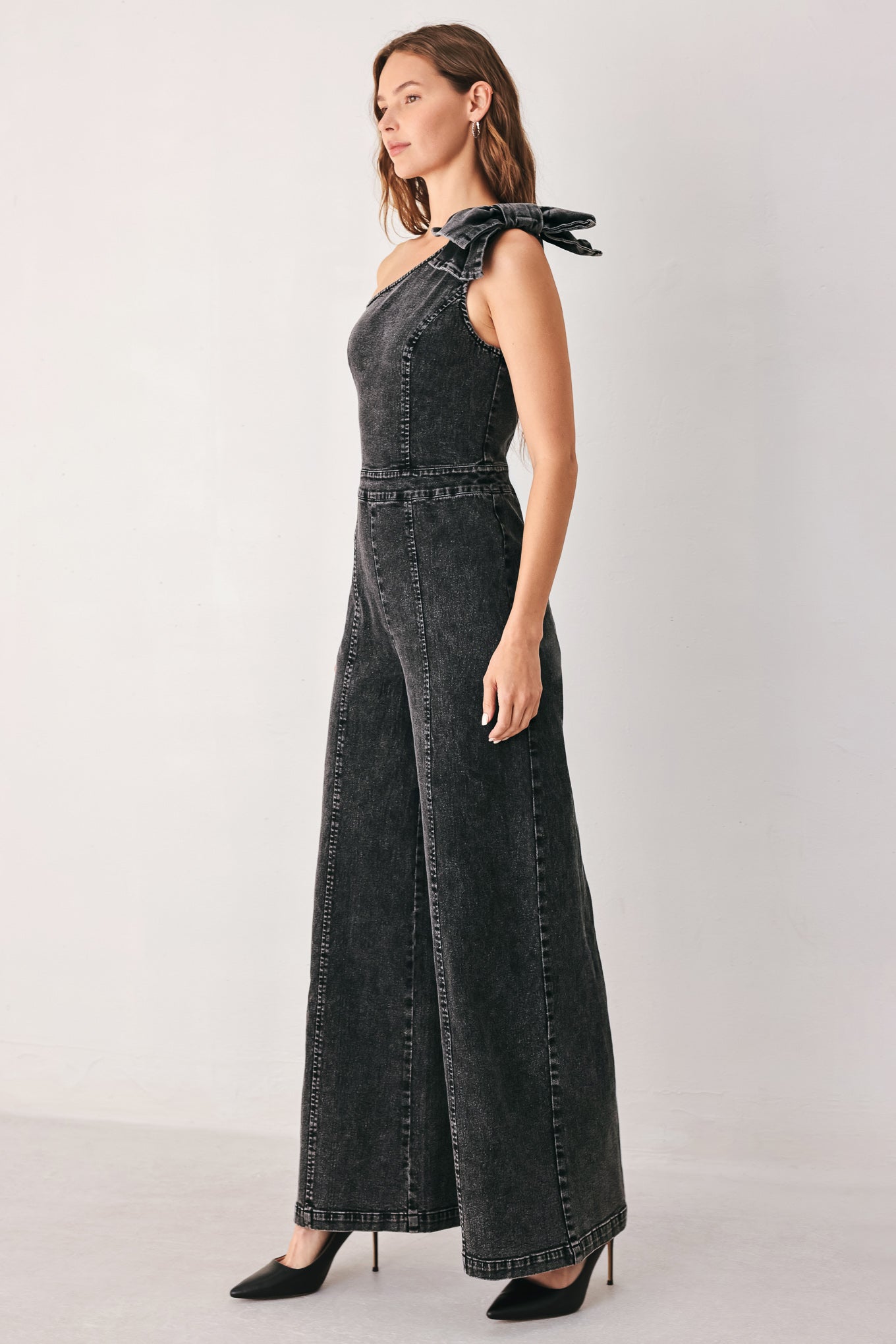 A woman wearing a dark gray washed denim one-shoulder jumpsuit with a wide-leg pant leg and a ruffled bow detail on the shoulder | FREEMARKET