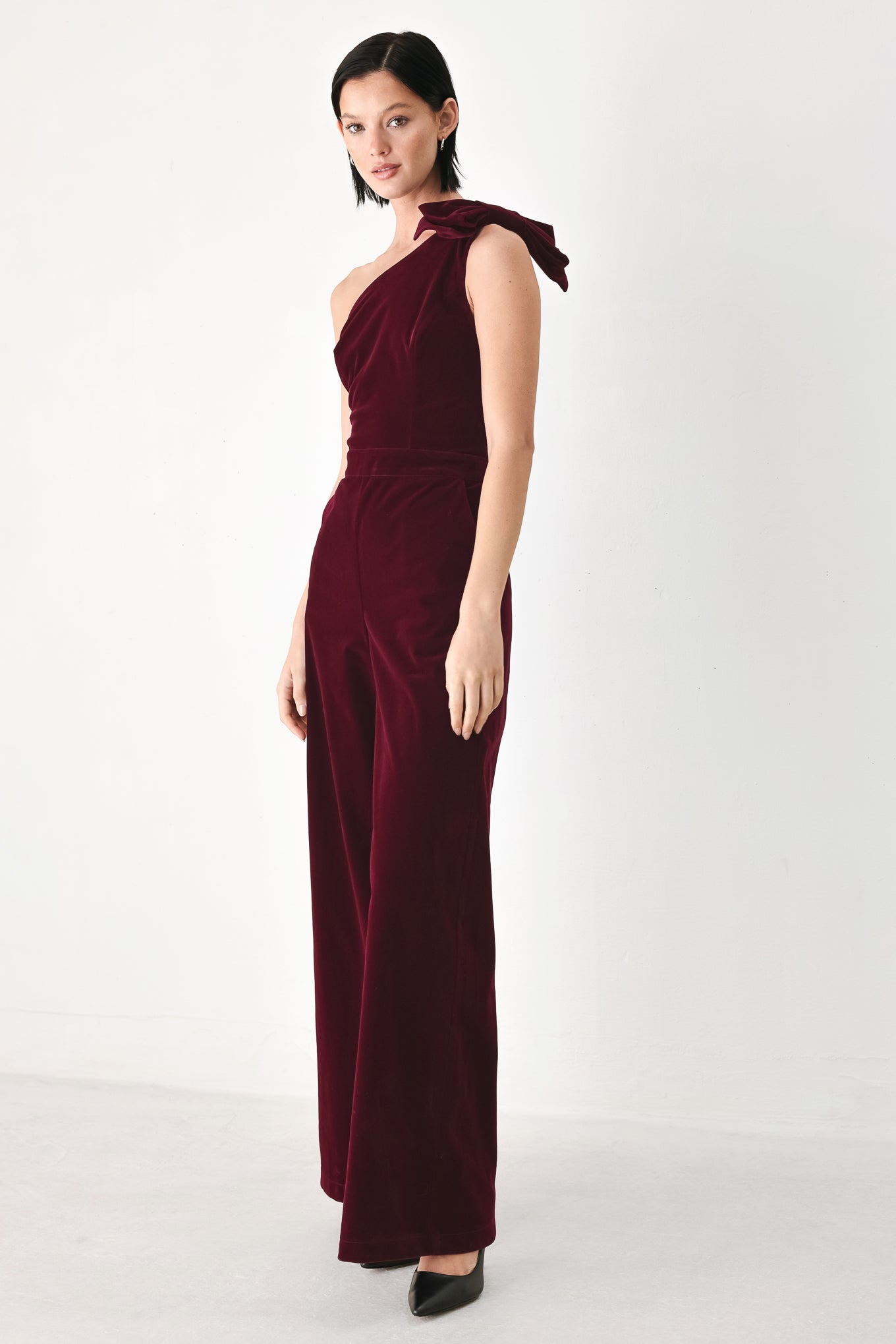 A woman wearing a deep burgundy velvet one-shoulder jumpsuit with a large bow detail on the shoulder | FREEMARKET