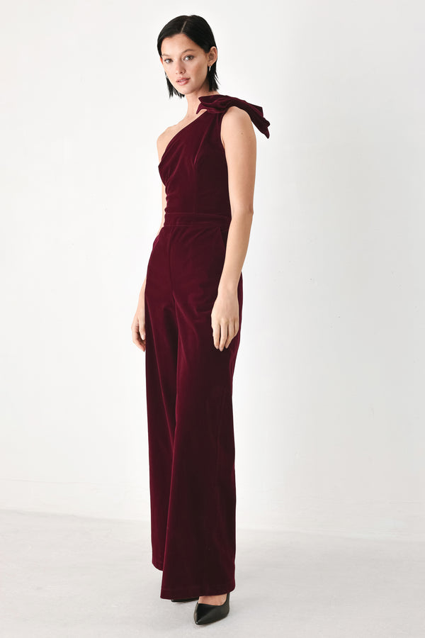 A woman wearing a deep burgundy velvet one-shoulder jumpsuit with a large bow detail on the shoulder | FREEMARKET