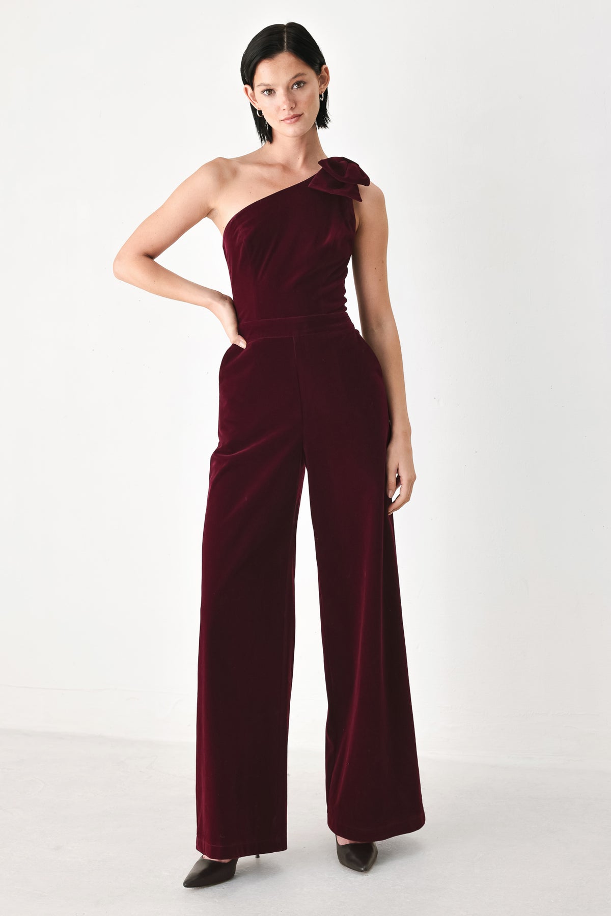 A woman wearing a deep burgundy velvet one-shoulder jumpsuit with a large bow detail on the shoulder | FREEMARKET