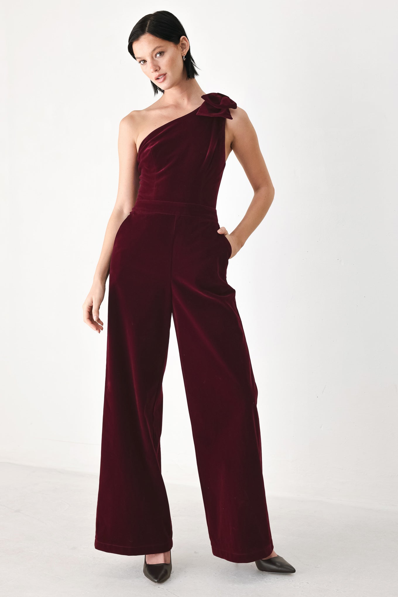 A woman wearing a deep burgundy velvet one-shoulder jumpsuit with a large bow detail on the shoulder | FREEMARKET