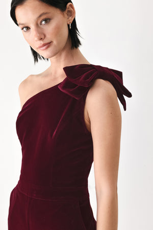 A woman wearing a deep burgundy velvet one-shoulder jumpsuit with a large bow detail on the shoulder | FREEMARKET