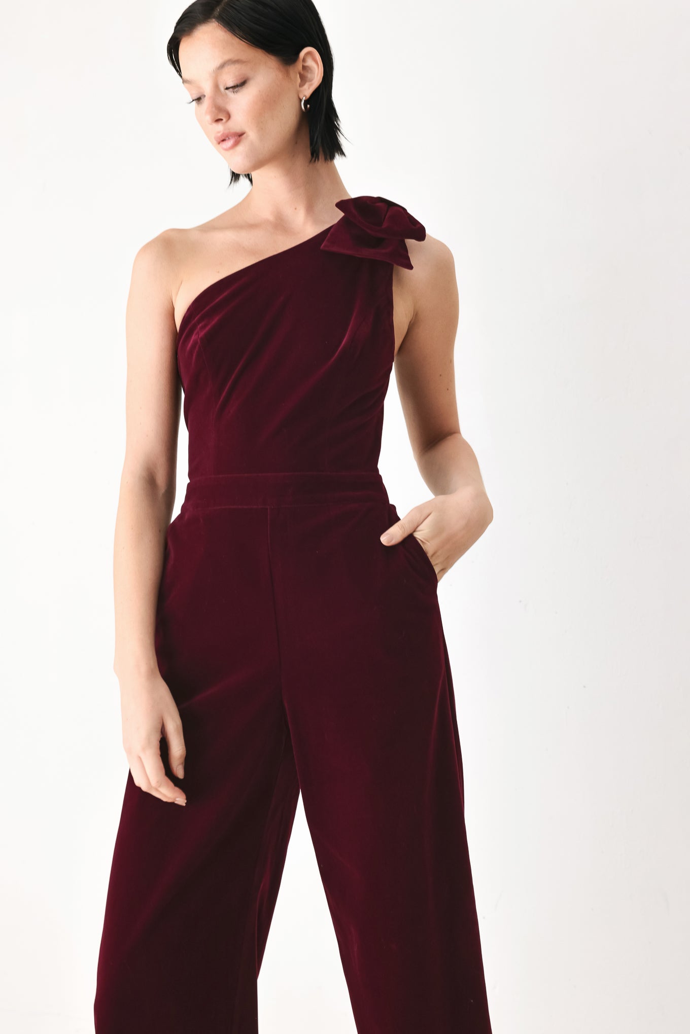 A woman wearing a deep burgundy velvet one-shoulder jumpsuit with a large bow detail on the shoulder | FREEMARKET