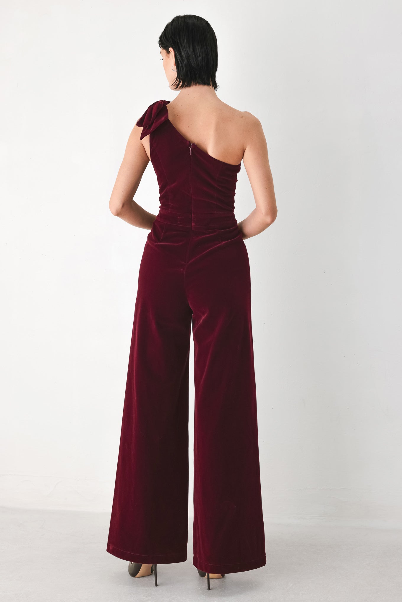 A woman wearing a deep burgundy velvet one-shoulder jumpsuit with a large bow detail on the shoulder | FREEMARKET