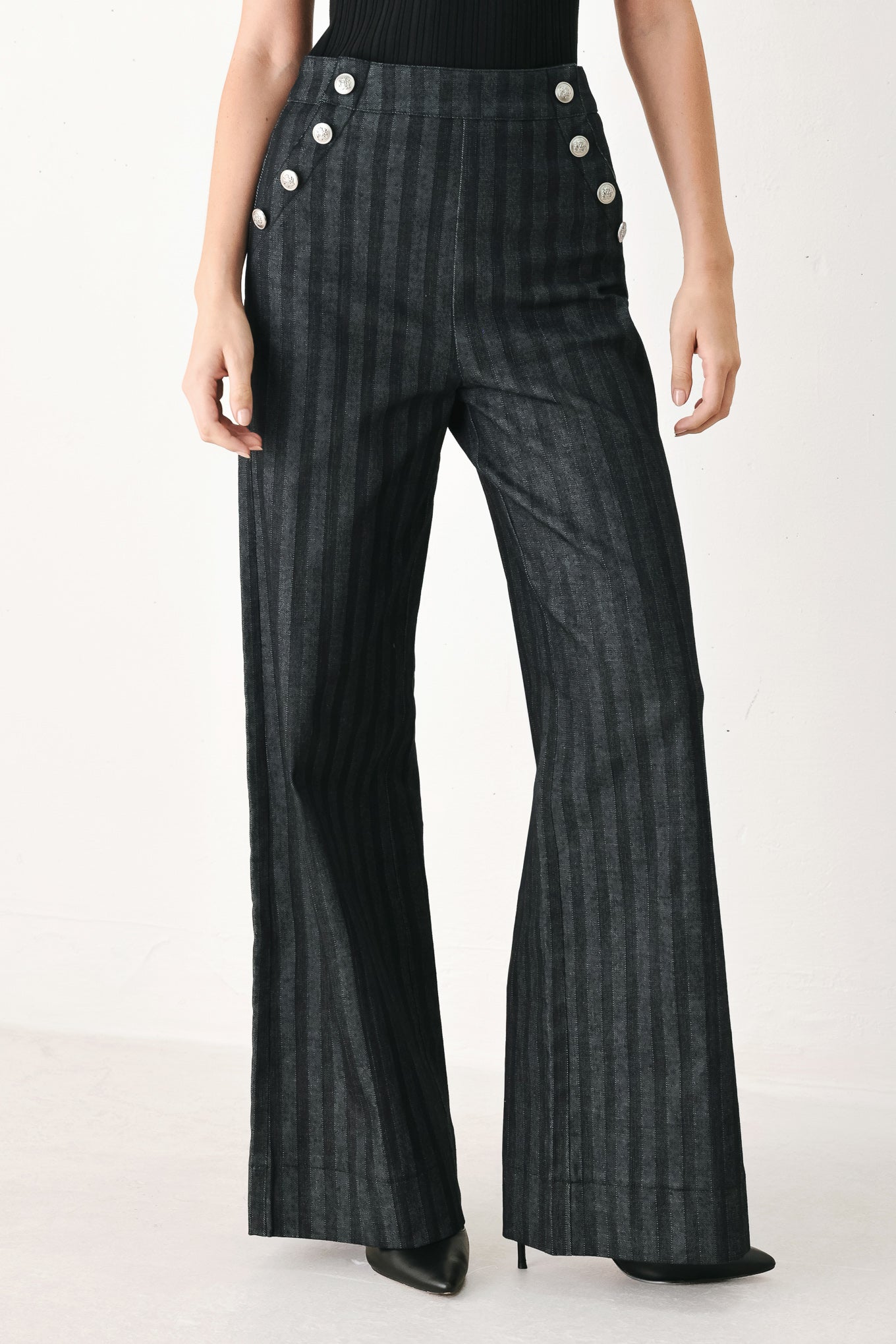 A model wearing a pair of high-waisted, wide-leg trousers in a dark gray and black pinstripe pattern, featuring decorative silver buttons on the front waistband | FREEMARKET