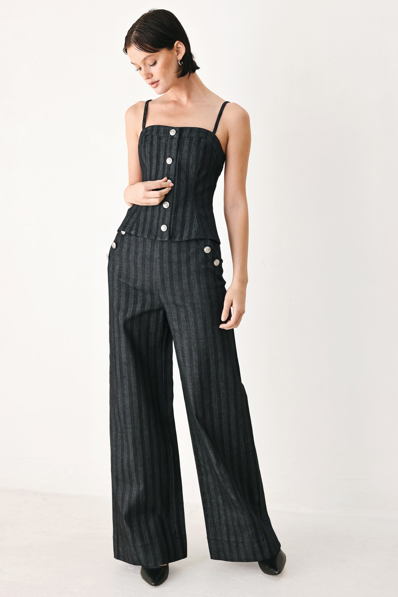 A model wearing a pair of high-waisted, wide-leg trousers in a dark gray and black pinstripe pattern, featuring decorative silver buttons on the front waistband | FREEMARKET