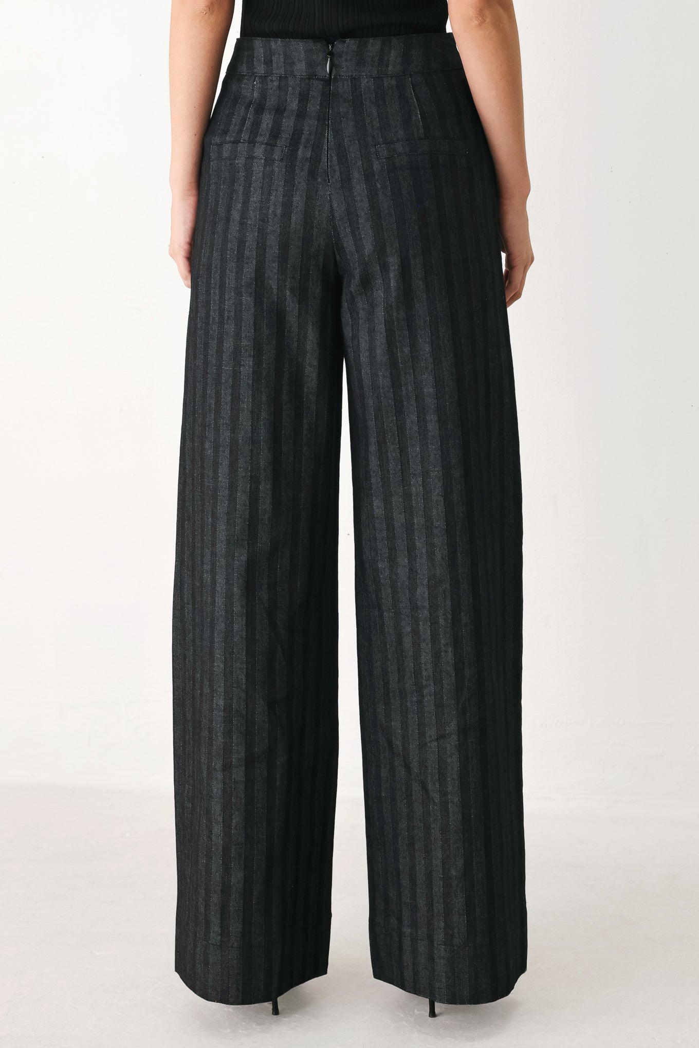 A model wearing a pair of high-waisted, wide-leg trousers in a dark gray and black pinstripe pattern, featuring decorative silver buttons on the front waistband | FREEMARKET