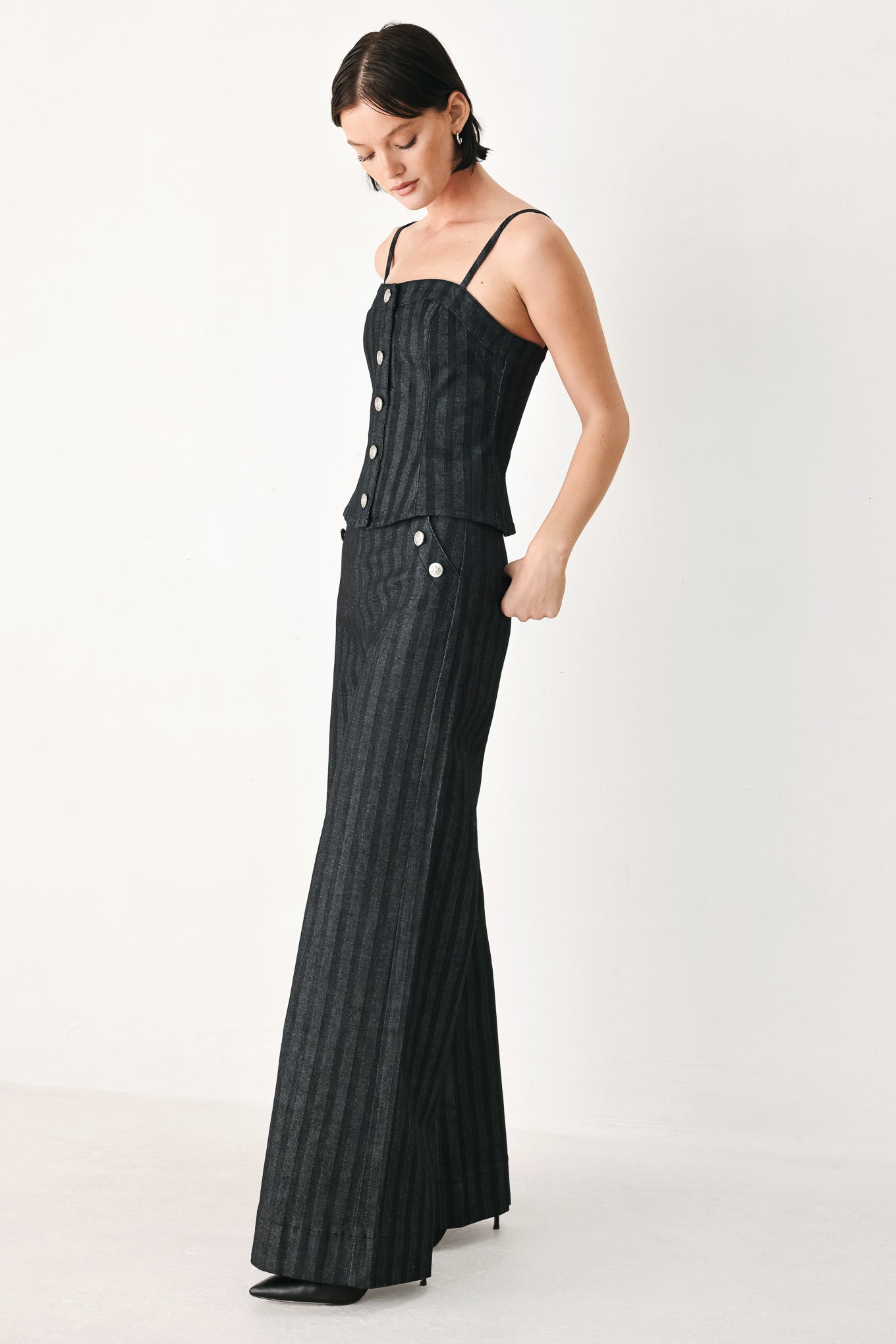 A model wearing a pair of high-waisted, wide-leg trousers in a dark gray and black pinstripe pattern, featuring decorative silver buttons on the front waistband | FREEMARKET