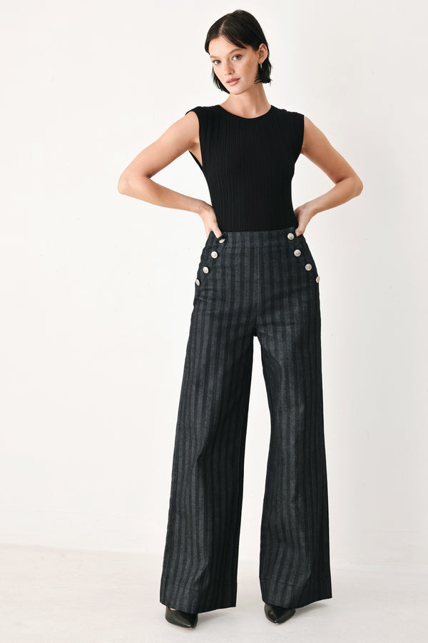 A model wearing a pair of high-waisted, wide-leg trousers in a dark gray and black pinstripe pattern, featuring decorative silver buttons on the front waistband | FREEMARKET