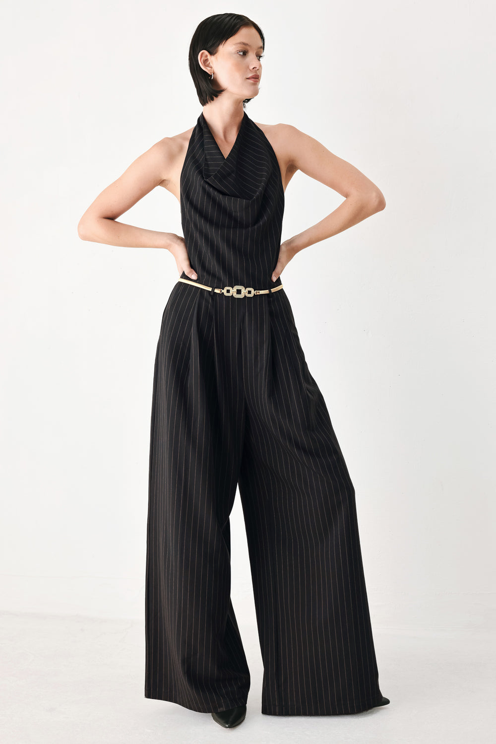 A model wearing a black pleated halter-neck jumpsuit with wide legs, pinstripes, and a gold chain belt | FREEMARKET