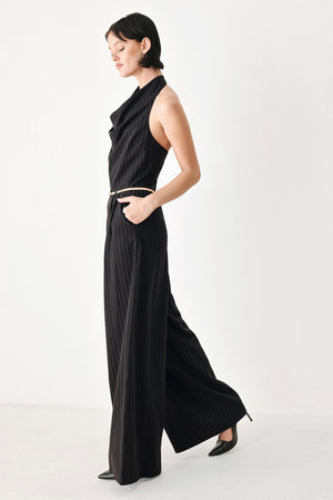 A model wearing a black pleated halter-neck jumpsuit with wide legs, pinstripes, and a gold chain belt | FREEMARKET
