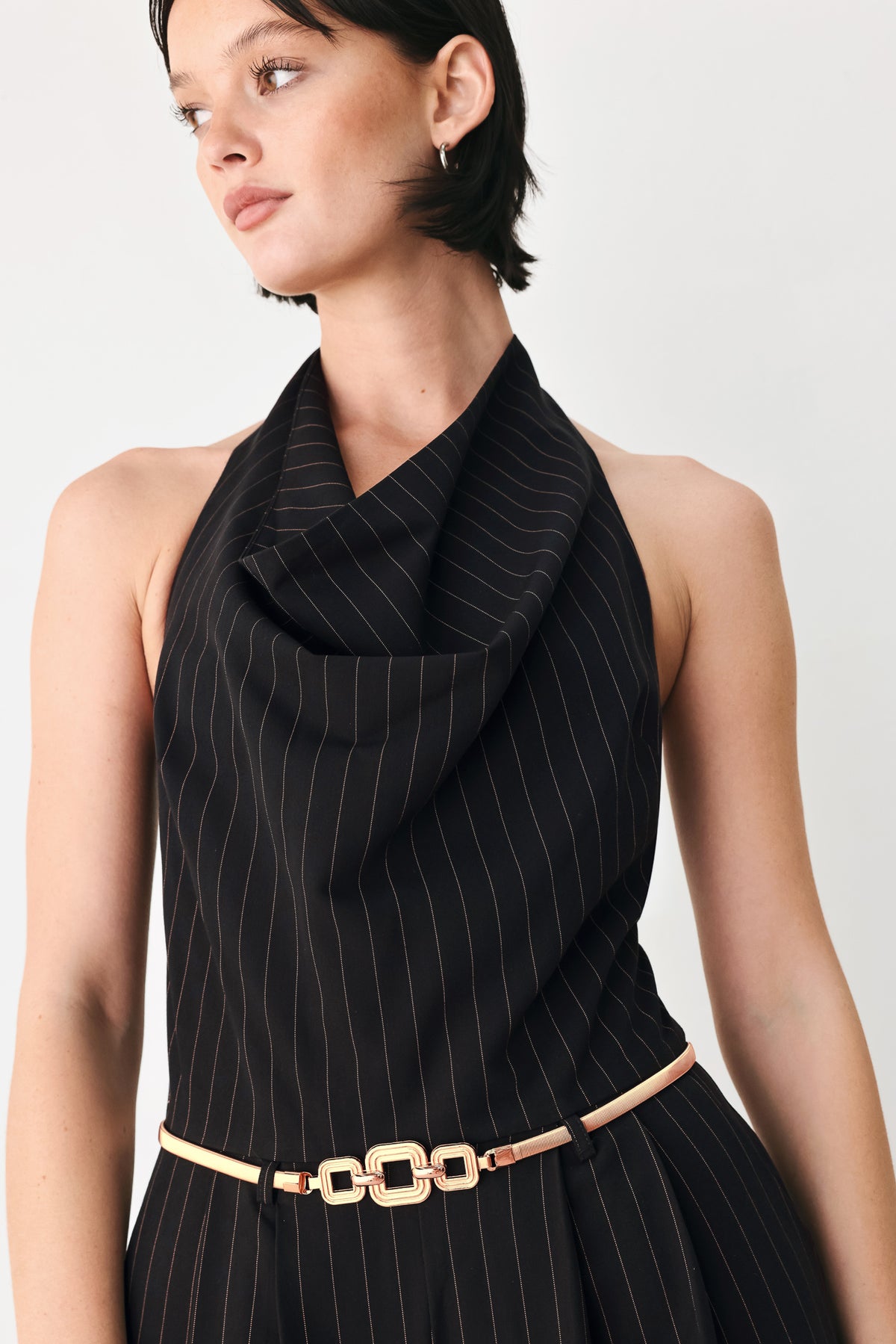 A model wearing a black pleated halter-neck jumpsuit with wide legs, pinstripes, and a gold chain belt | FREEMARKET