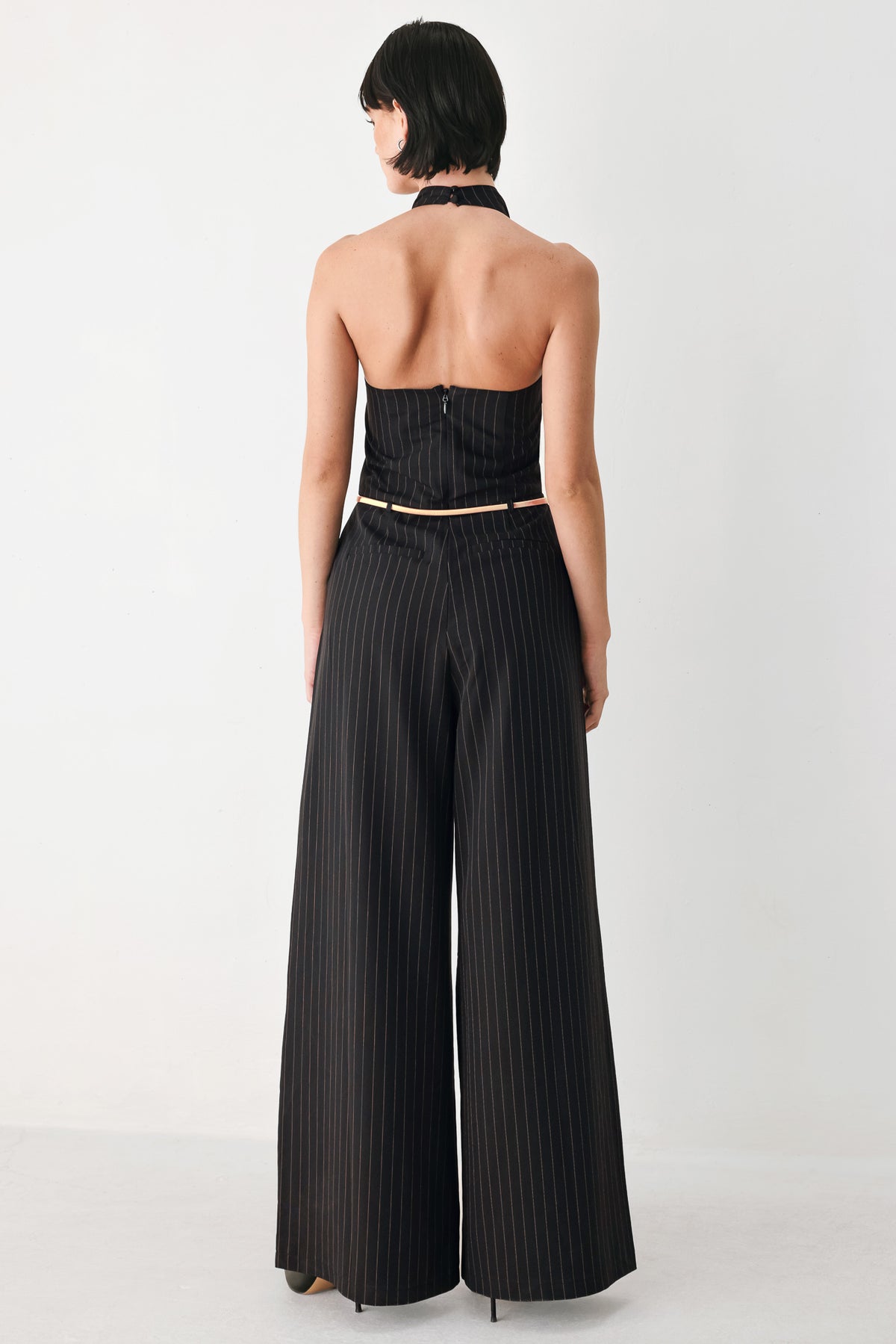 A model wearing a black pleated halter-neck jumpsuit with wide legs, pinstripes, and a gold chain belt | FREEMARKET