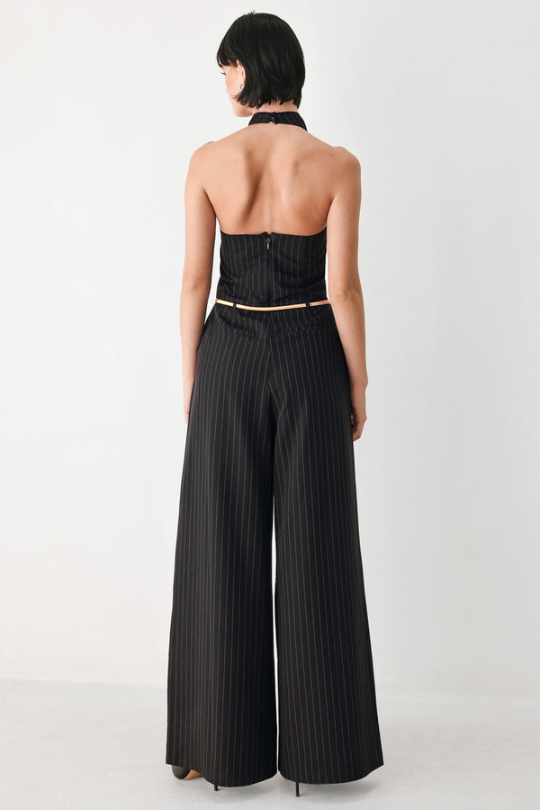A model wearing a black pleated halter-neck jumpsuit with wide legs, pinstripes, and a gold chain belt | FREEMARKET