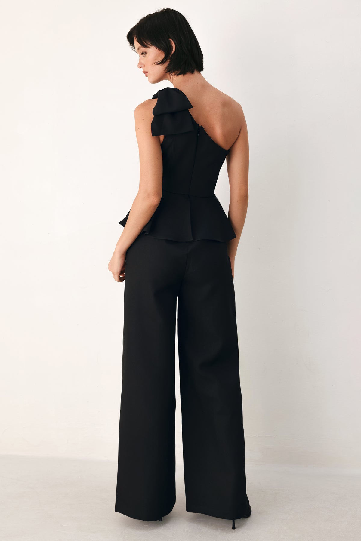 A model wearing a black one-shoulder jumpsuit with a ruffled bodice and wide-leg trousers | FREEMARKET