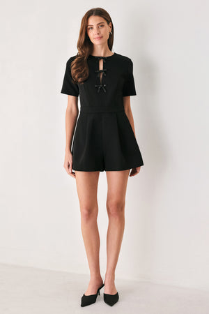Woman wearing a black short sleeve romper with front keyhole and small embellished bows and pockets | FREEMARKET