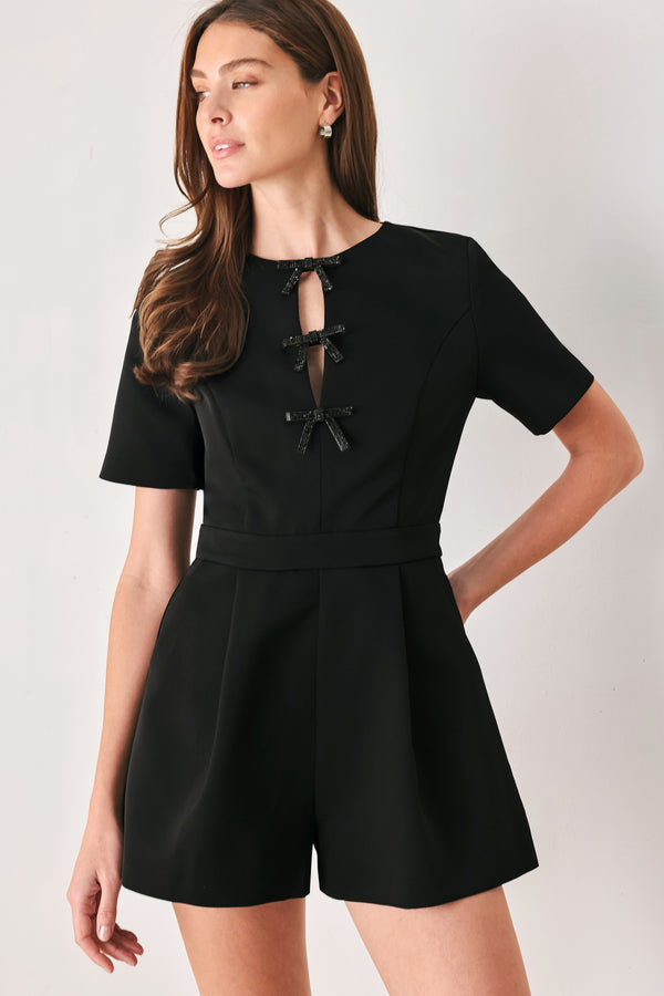 Woman wearing a black short sleeve romper with front keyhole and small embellished bows and pockets | FREEMARKET