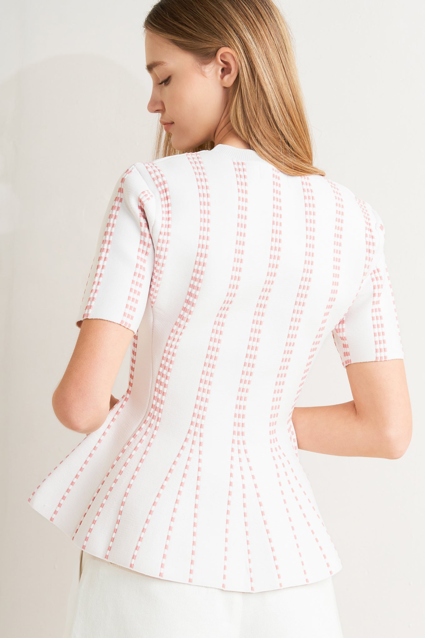 Woman wearing a pink blouse with white stitching against a neutral background AND A jacquard sweater top featuring round neckline, short sleeve and peplum FLYING TOMATO