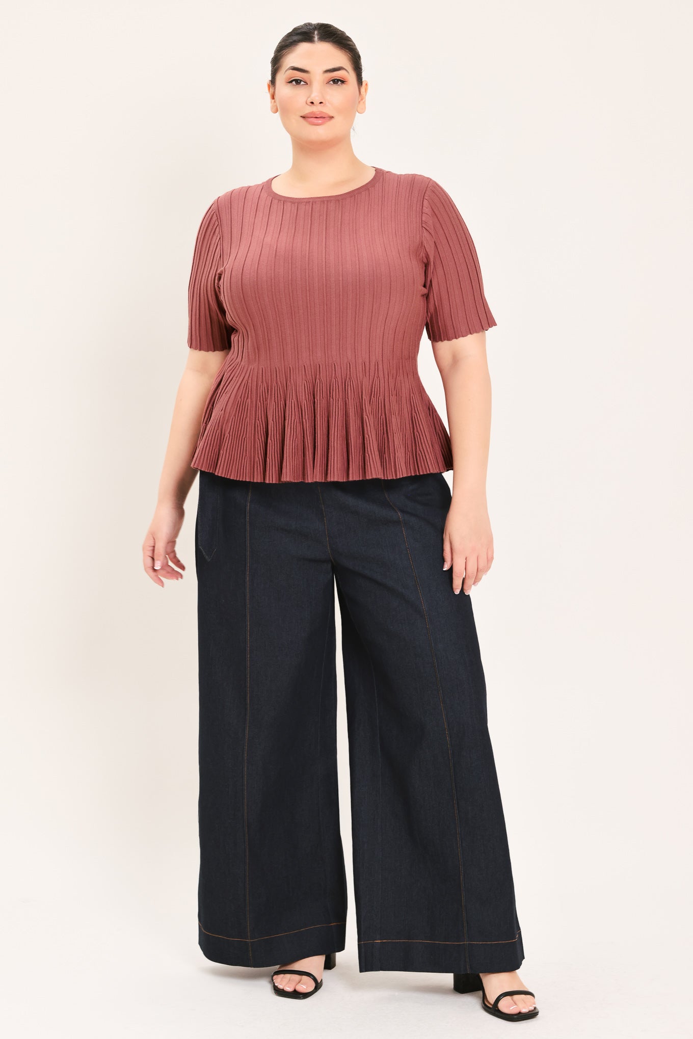 A woman modeling a short-sleeve, crew-neck top with a pleated texture and peplum hem in a dusty rose color | Plus Curve Flying Tomato