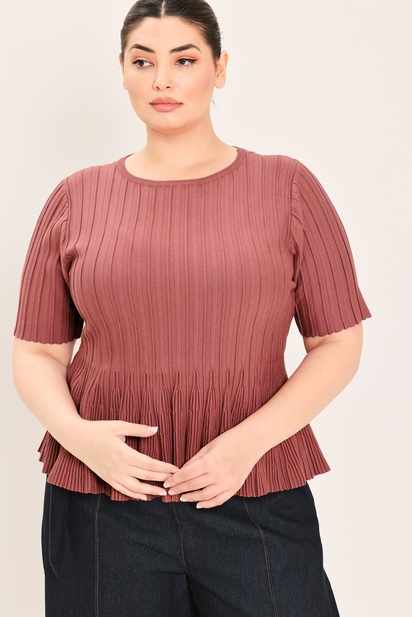 A woman modeling a short-sleeve, crew-neck top with a pleated texture and peplum hem in a dusty rose color | Plus Curve Flying Tomato
