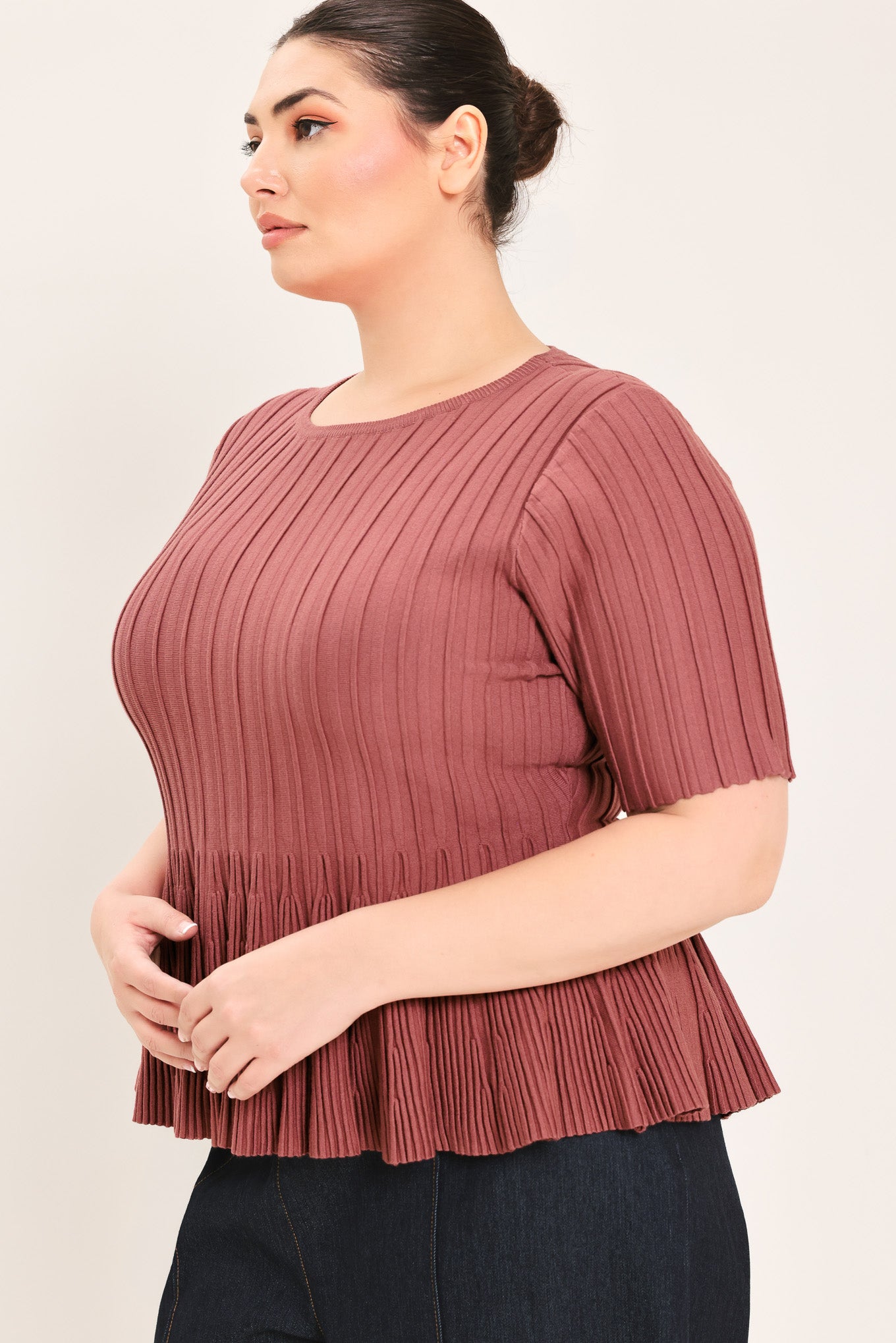 A woman modeling a short-sleeve, crew-neck top with a pleated texture and peplum hem in a dusty rose color | Plus Curve Flying Tomato