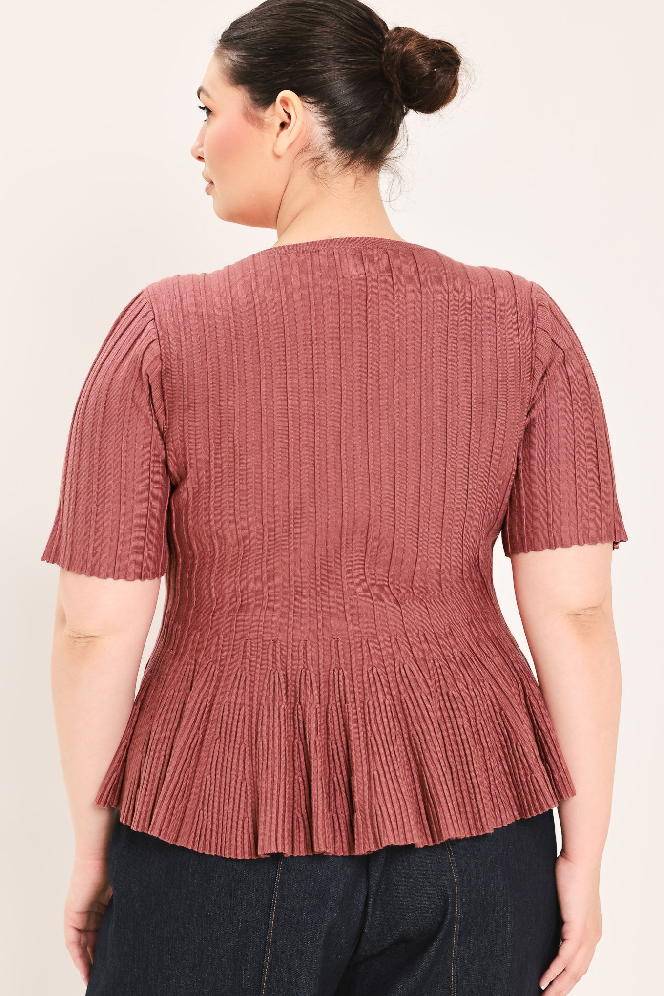 A woman modeling a short-sleeve, crew-neck top with a pleated texture and peplum hem in a dusty rose color | Plus Curve Flying Tomato