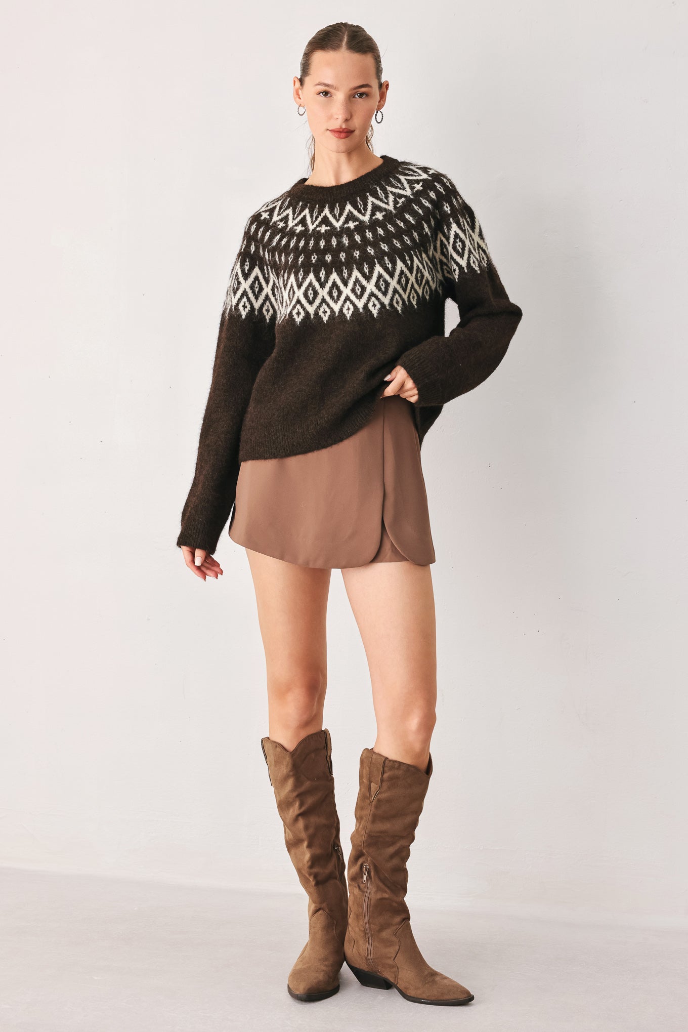 A model stands against a light gray studio background wearing a dark brown knit sweater with a white geometric pattern across the chest | FREEMARKET