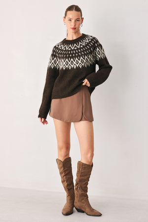 A model stands against a light gray studio background wearing a dark brown knit sweater with a white geometric pattern across the chest | FREEMARKET