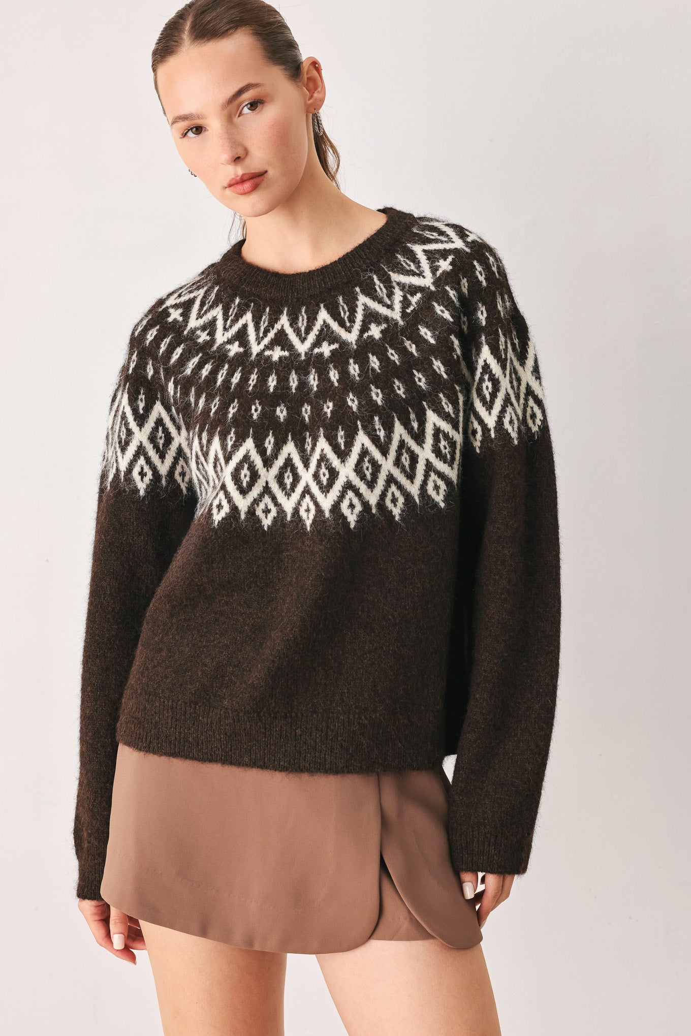 A model stands against a light gray studio background wearing a dark brown knit sweater with a white geometric pattern across the chest | FREEMARKET