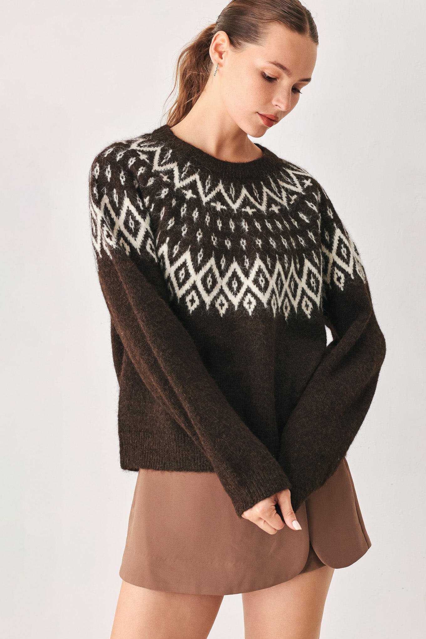 A model stands against a light gray studio background wearing a dark brown knit sweater with a white geometric pattern across the chest | FREEMARKET