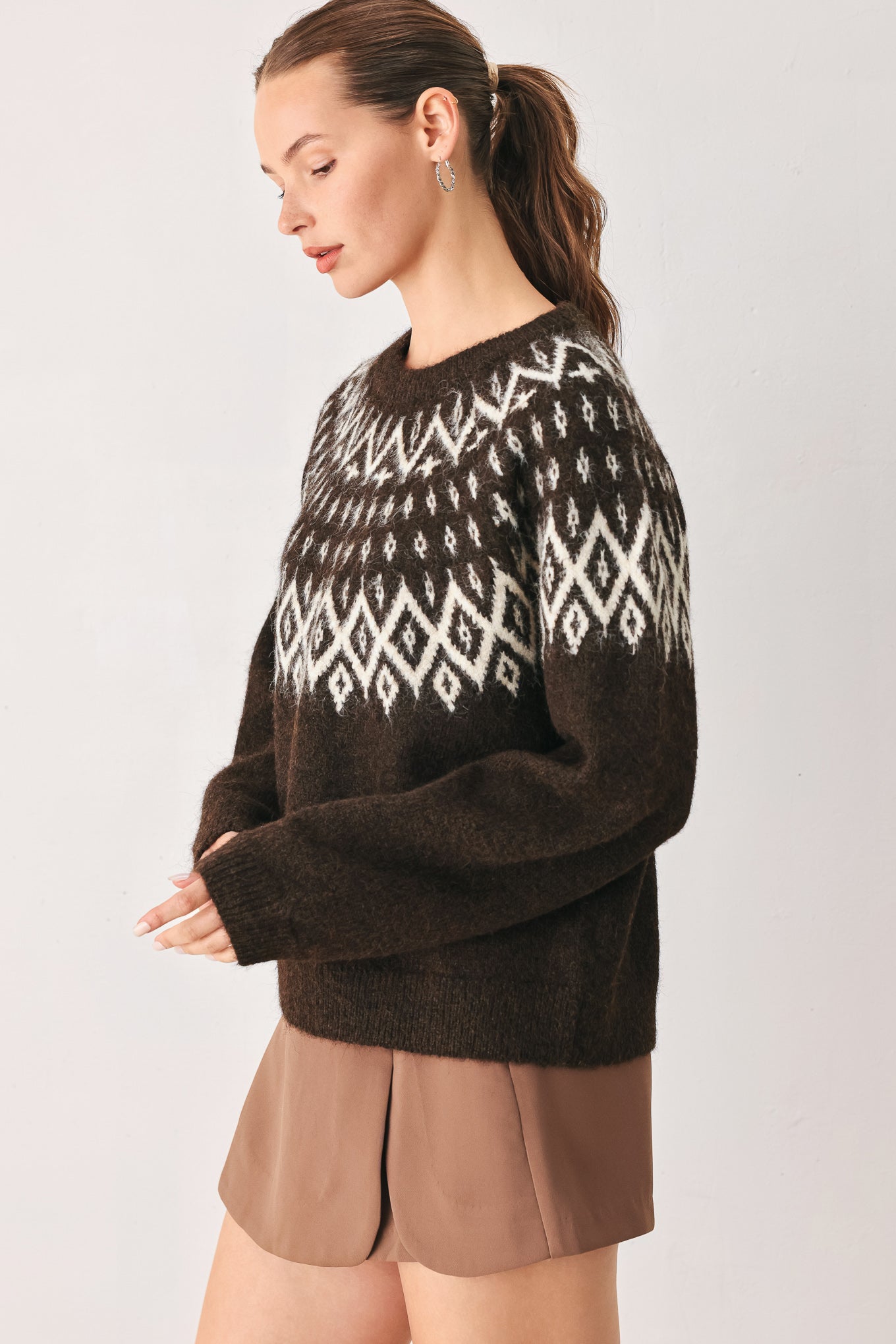A model stands against a light gray studio background wearing a dark brown knit sweater with a white geometric pattern across the chest | FREEMARKET