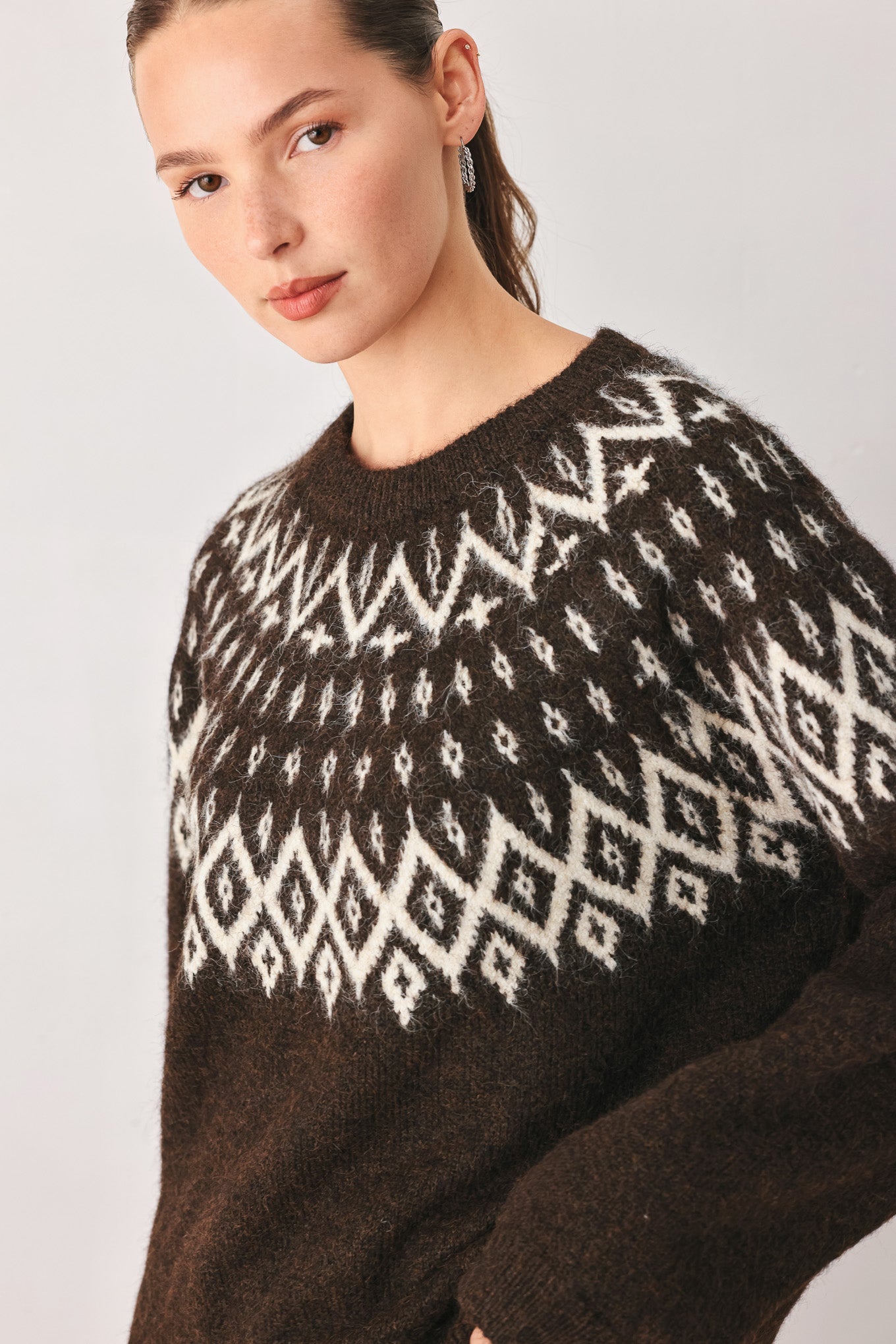 A model stands against a light gray studio background wearing a dark brown knit sweater with a white geometric pattern across the chest | FREEMARKET
