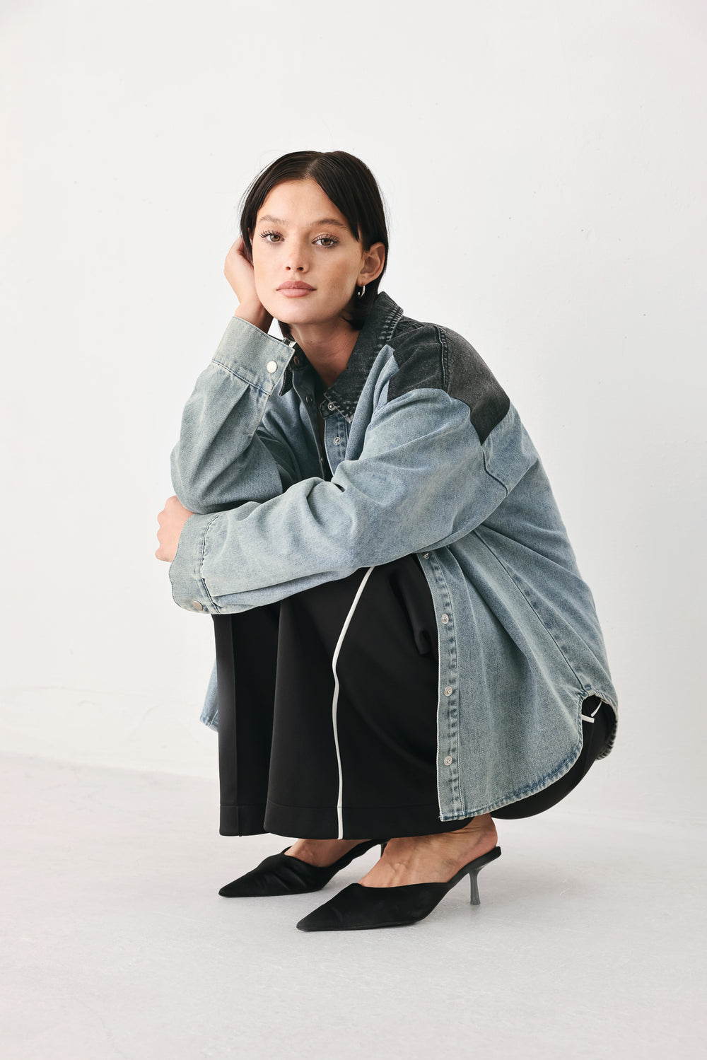 A model wearing a light-wash denim button-up shirt jacket with black contrast panels on the shoulders and collar, styled over a black top and black trousers against a light gray studio background.
