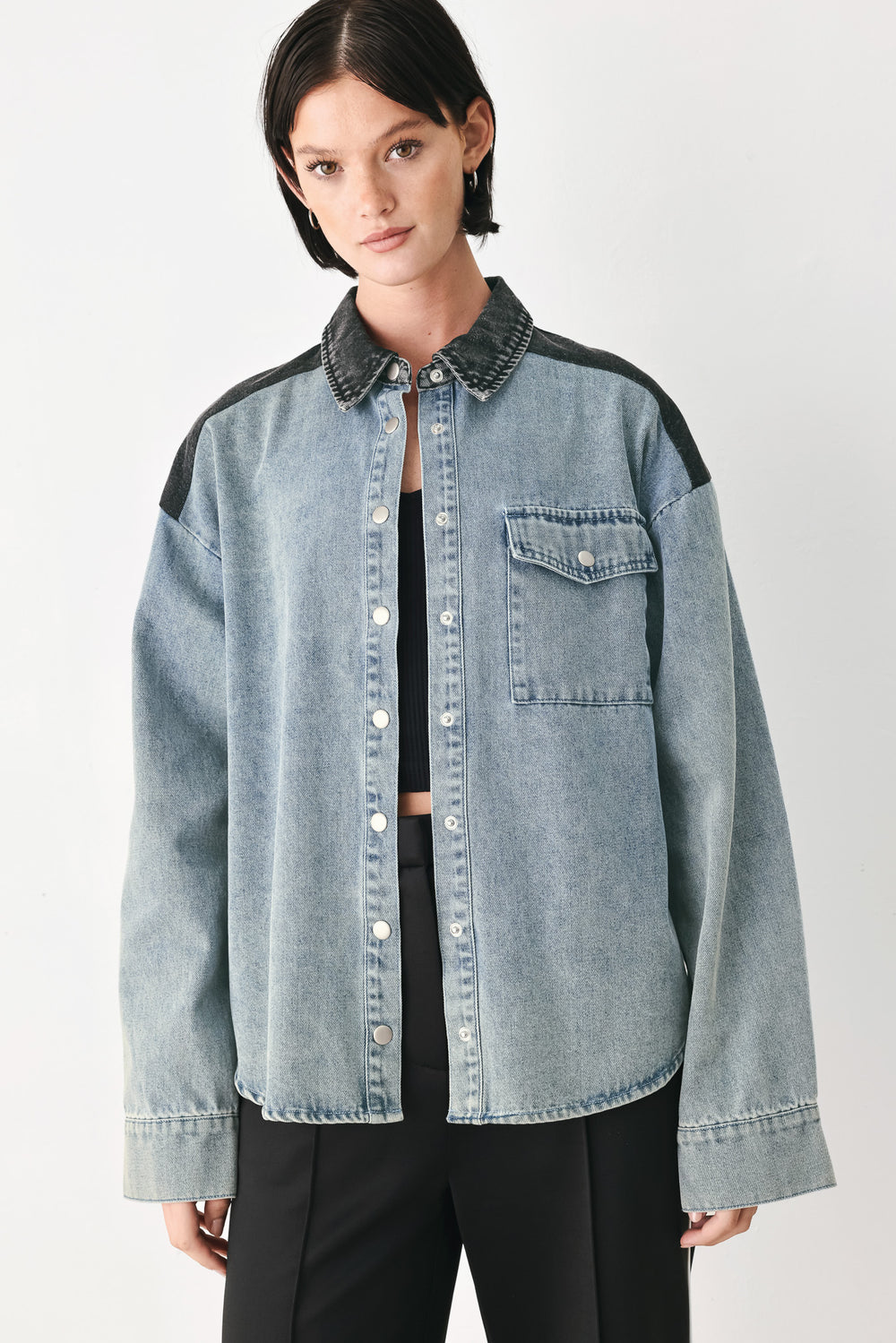 A model wearing a light-wash denim button-up shirt jacket with black contrast panels on the shoulders and collar, styled over a black top and black trousers against a light gray studio background.