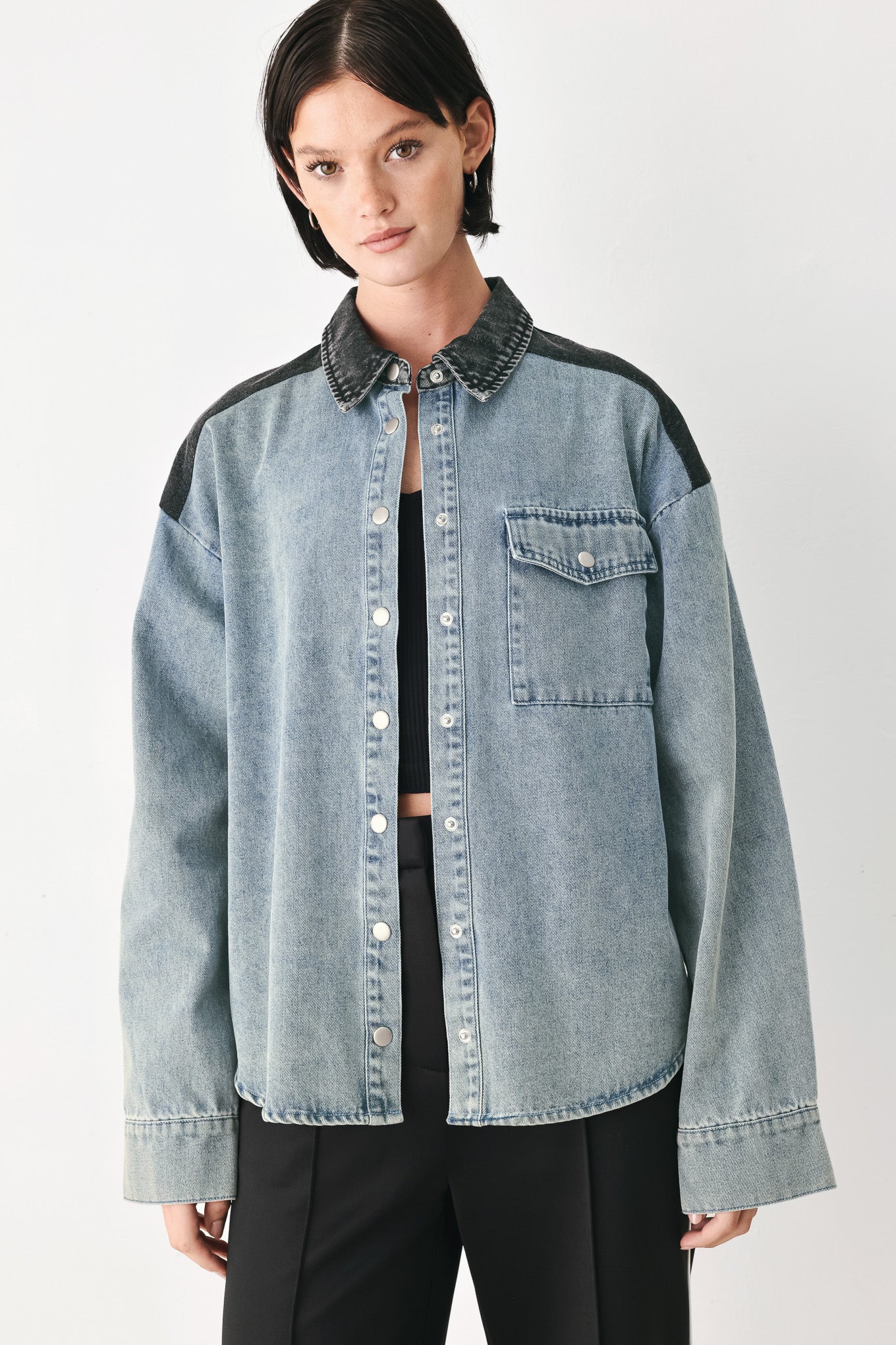 A model wearing a light-wash denim button-up shirt jacket with black contrast panels on the shoulders and collar, styled over a black top and black trousers against a light gray studio background.