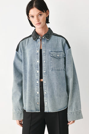 A model wearing a light-wash denim button-up shirt jacket with black contrast panels on the shoulders and collar, styled over a black top and black trousers against a light gray studio background.